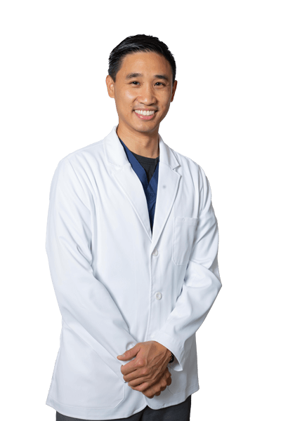 The image shows a man wearing a white lab coat, standing with his hands together in front of him, and smiling at the camera. He appears to be a medical professional, possibly a doctor or a nurse.