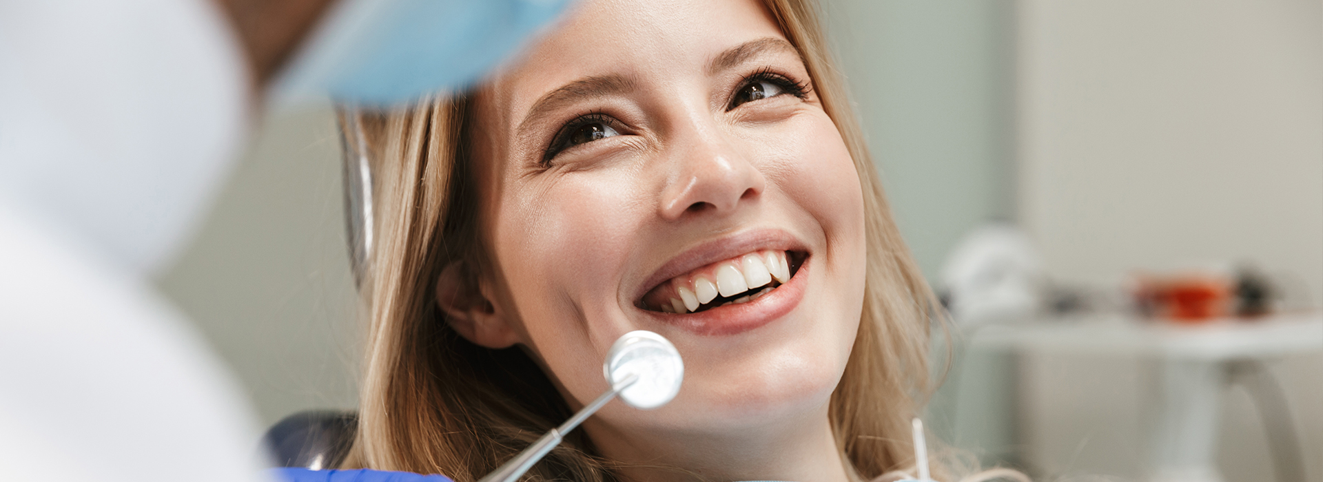 The image shows a smiling woman in a dental setting, receiving attention from a dental professional holding a mirror.